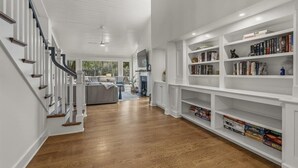 Un pasillo amplio con una escalera, una sala de estar con sofá, y una estantería llena de libros.