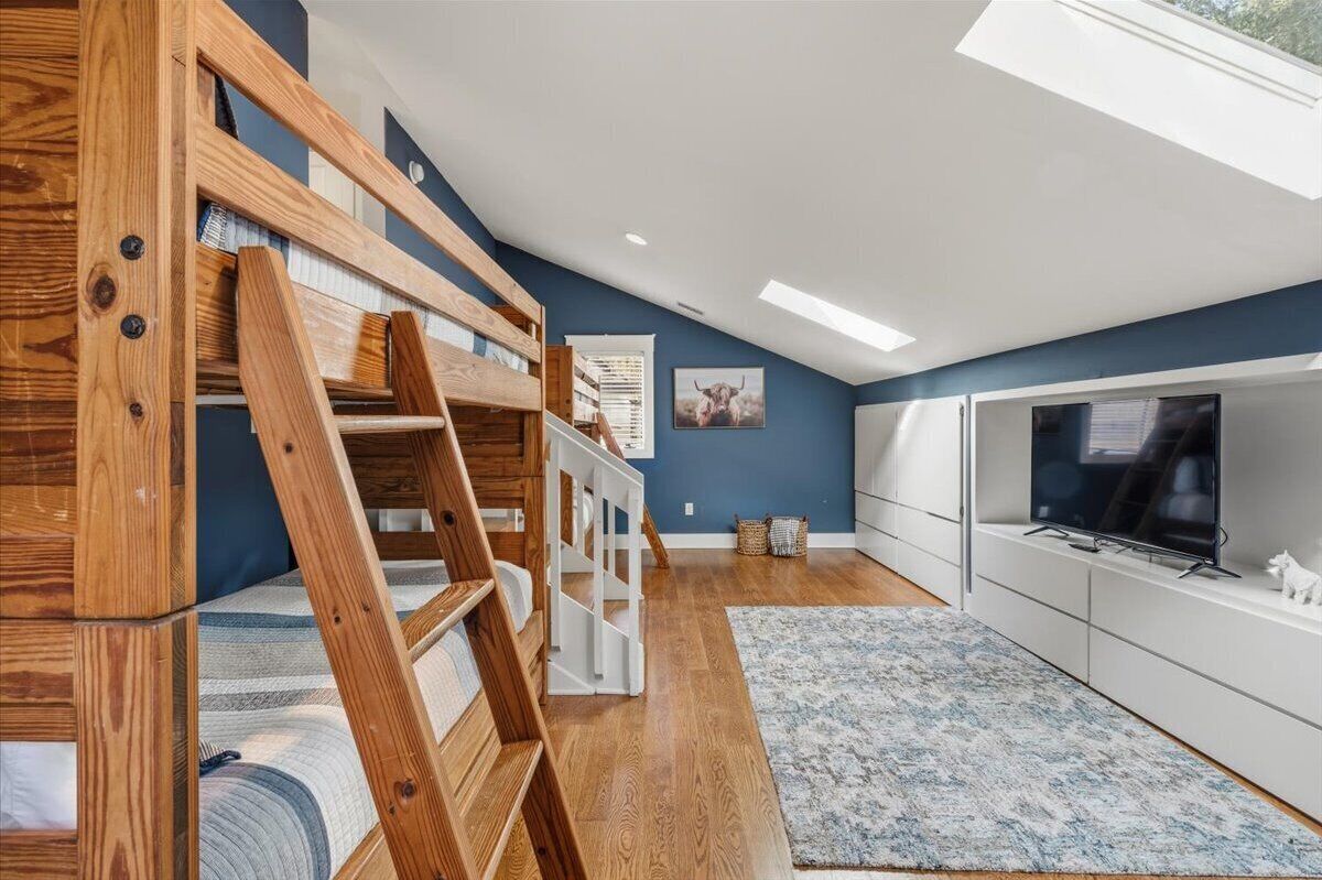 A loft bedroom with a wooden bunk bed, a skylight, a flat-screen TV, and a large window.