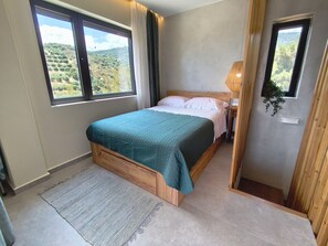 Ein Schlafzimmer mit einem Bett, einem Fenster, einer Nachttischlampe und Blick auf eine hügelige Landschaft.