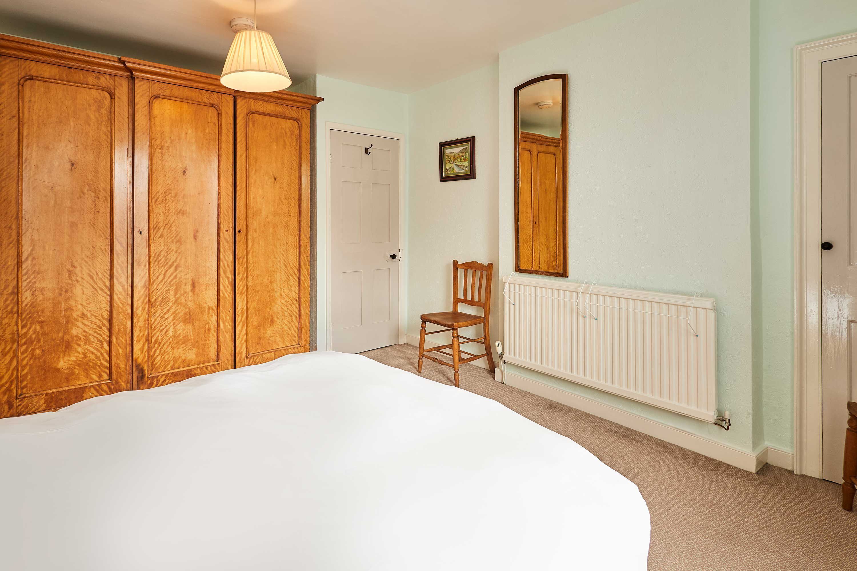 A bedroom with a large bed, wooden wardrobe, a chair, a radiator, and a door.
