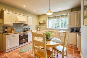 A kitchen with white cabinets, a wooden dining table with chairs, a hanging light fixture, and a window with a view of greenery. - Host Stay Tree Tops (Darlington)