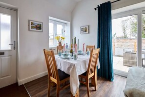 Uma área de jantar com mesa posta para quatro pessoas, cadeiras de madeira, toalha de mesa branca e vista através de uma porta de vidro para uma área externa.