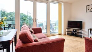 Uma sala de estar com sofá, televisão em móvel, mesa de jantar com cadeiras e grandes janelas.