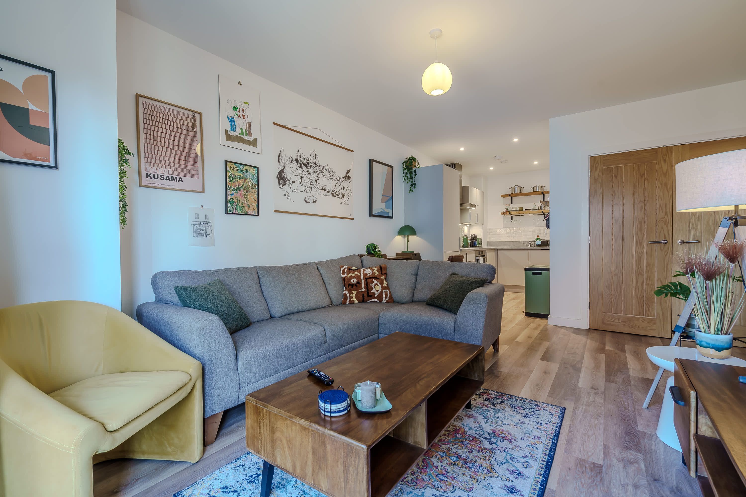 Un salon moderne avec un canapé, une table basse en bois et une table à manger. On peut y voir des tableaux encadrés au mur, une lampe et une plante.