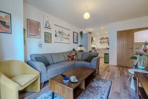 Un salon moderne avec un canapé, une table basse en bois et une table à manger. On peut y voir des tableaux encadrés au mur, une lampe et une plante.