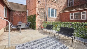 A courtyard with a brick building, a metal bench, a table with chairs, and a patterned rug. - Host Stay Paylors View Cottage (Whitby)