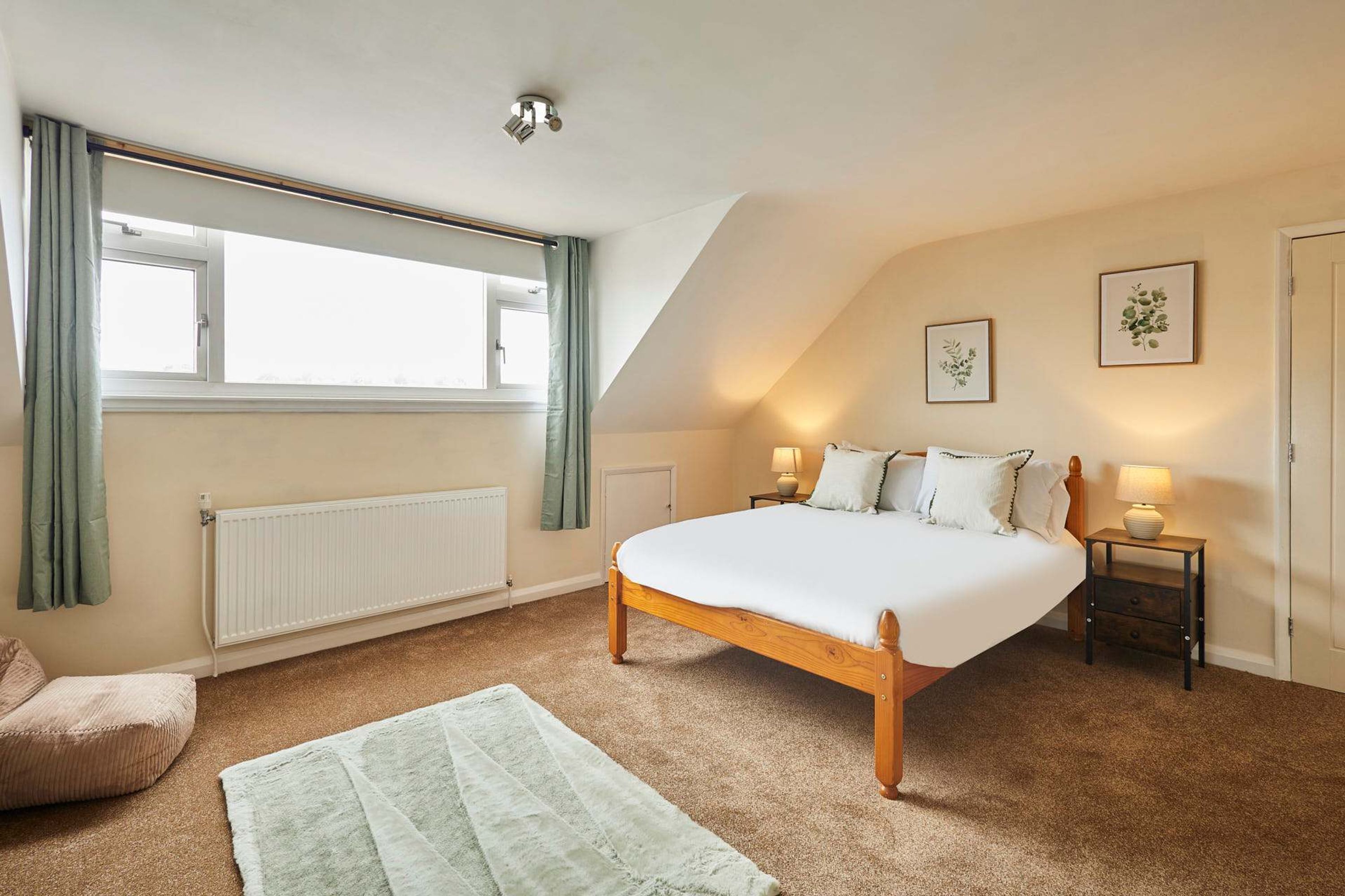 A bedroom with a large bed, a radiator, a window with curtains, a bedside table with a lamp, and a wall-mounted picture.