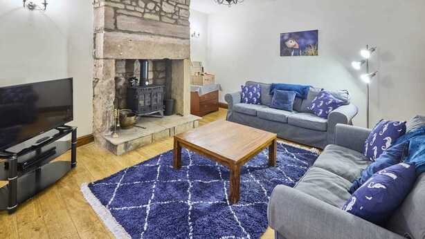 A living room with a stone fireplace, a sofa, a coffee table, and a flat-screen TV. - Host Stay Puffin Cottage (Seahouses)