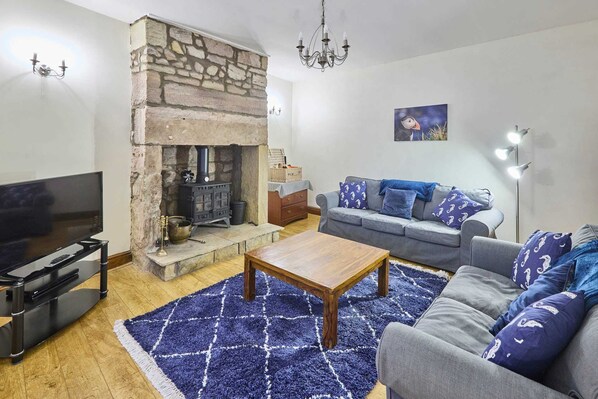 A living room with a stone fireplace, a sofa, a coffee table, and a flat-screen TV. - Host Stay Puffin Cottage (Seahouses)