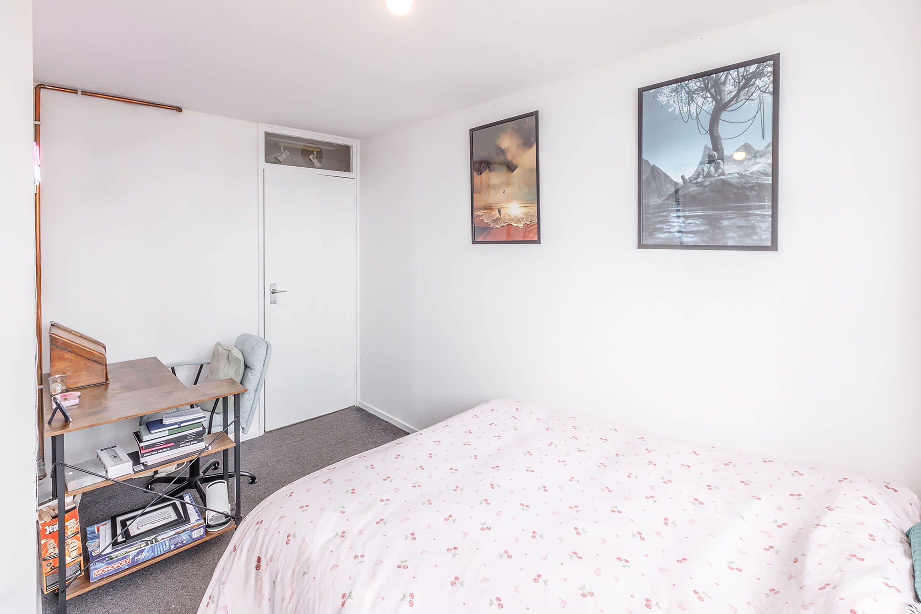 A bedroom with a bed, a desk with books, a chair, and two framed pictures on the wall.
