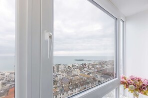 Ein Fenster mit Blick auf eine Küstenstadt, ein Schiff in der Ferne und eine Vase mit Blumen auf der Fensterbank.