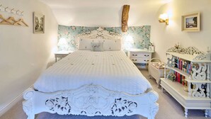 A bedroom with a white ornate bed, a floral wallpaper backdrop, a wooden beam, a bedside table with a lamp, and a bookshelf. - Host Stay Wistermead Cottage (Winkleigh)