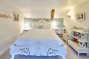 Un dormitorio con una cama blanca ornamentada, un fondo de papel tapiz floral, una viga de madera, una mesita de noche con lámpara y una estantería.