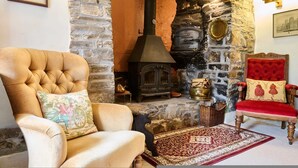 A traditional stone fireplace with a wood-burning stove, a red upholstered chair, and a cushioned armchair. - Host Stay Wistermead Cottage (Winkleigh)