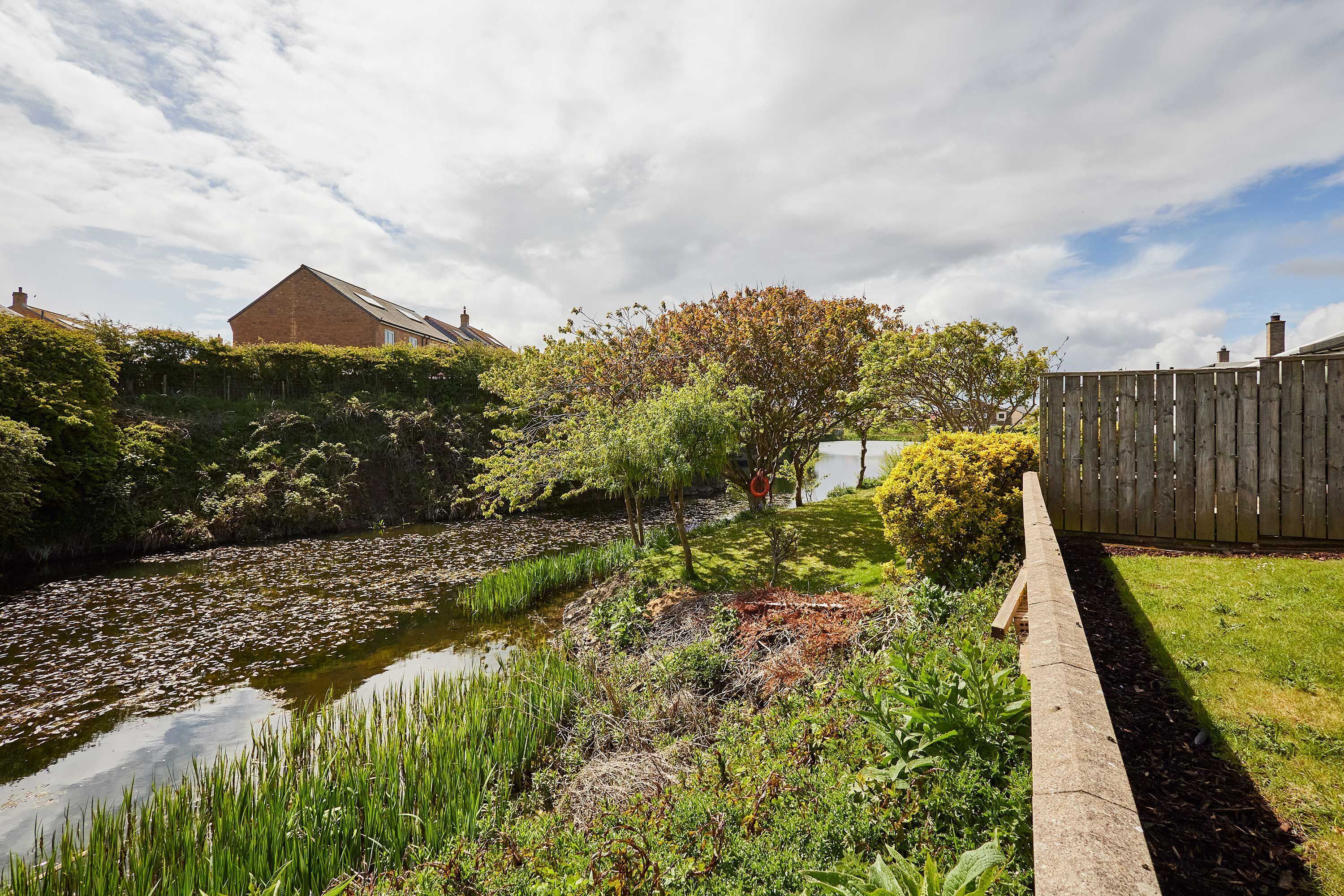 Um jardim com lago, cerca de madeira e uma casa ao fundo.