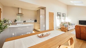 A modern kitchen with a central island, a dining area with a wooden table, and a living room with a sofa and TV. - Host Stay Stille House (Chathill)