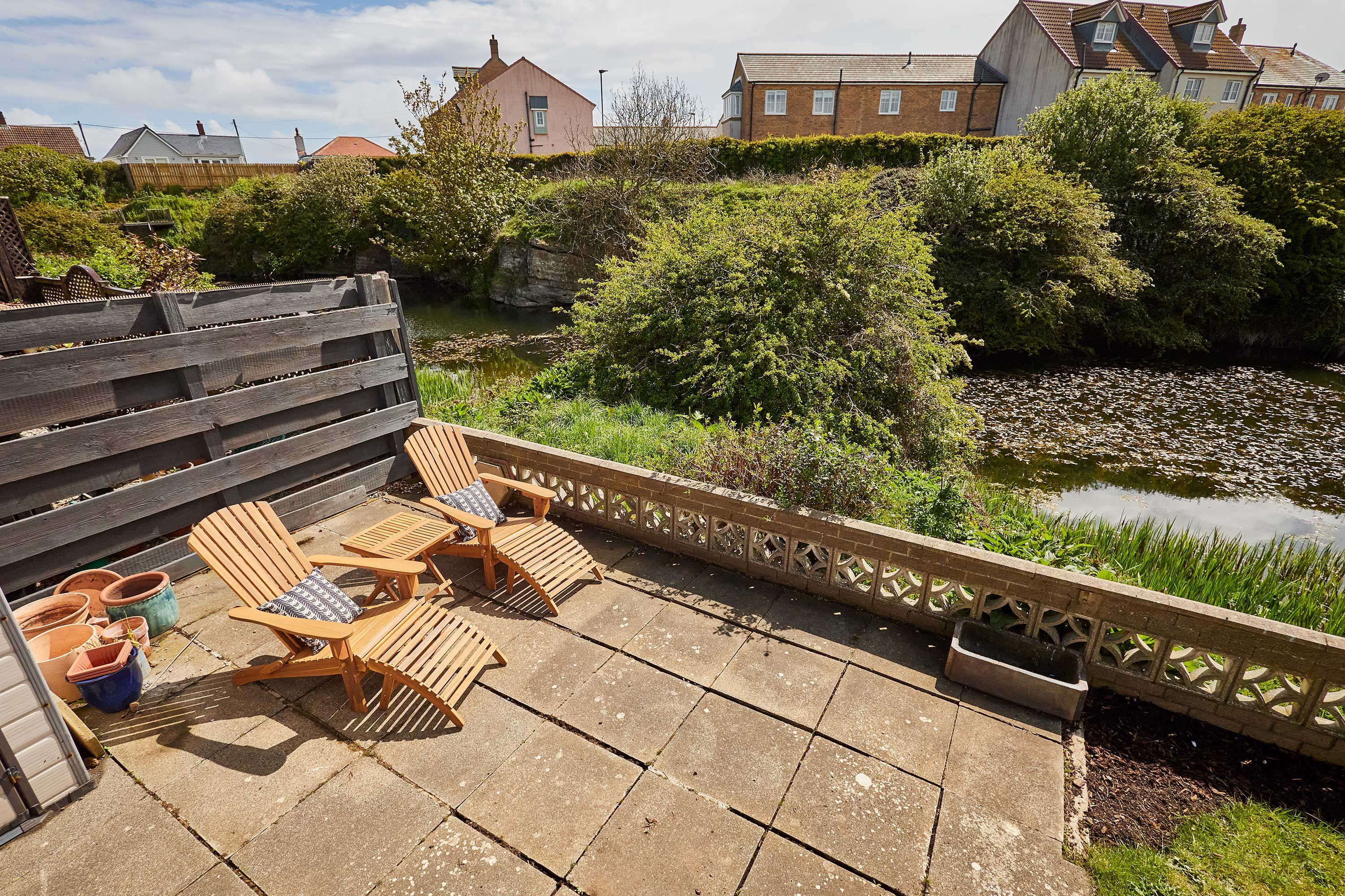 Une terrasse avec des chaises et une table en bois, un petit étang et une clôture en bois.