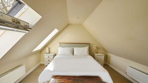 Quarto com teto inclinado, cama com roupa de cama branca, duas mesas de cabeceira e um banco de madeira.
