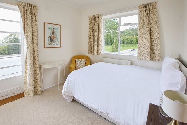 A bedroom with a large bed, a small table, a chair, and a window with curtains. - Host Stay Joldwynds (Dorking)