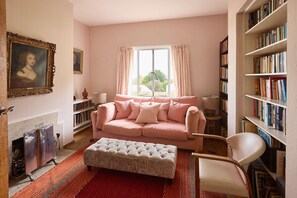 A living room with a pink sofa, a white ottoman, a fireplace, and a bookshelf filled with books. - Host Stay Joldwynds (Dorking)