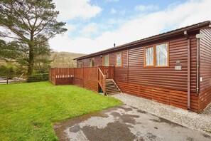 A wooden cabin with a staircase leading to a deck, surrounded by a grassy yard and a large tree. - Host Stay Pine View (Windermere)