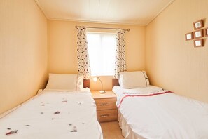 A bedroom with two single beds, a nightstand with a lamp, a window with curtains, and a wall with framed pictures. - Host Stay Pine View (Windermere)