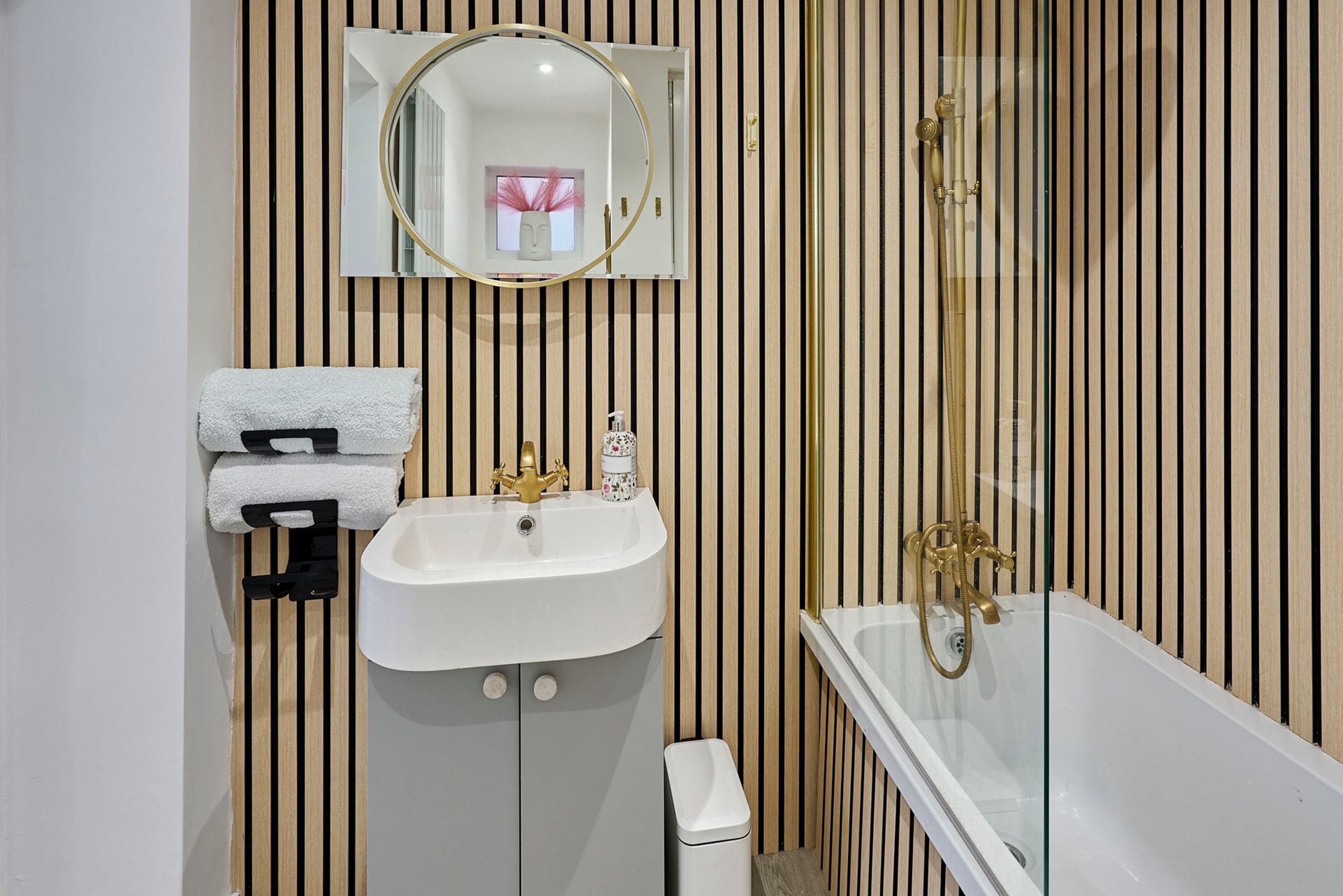 A bathroom with a white sink, a mirror, a bathtub with gold fixtures, and a striped wall.