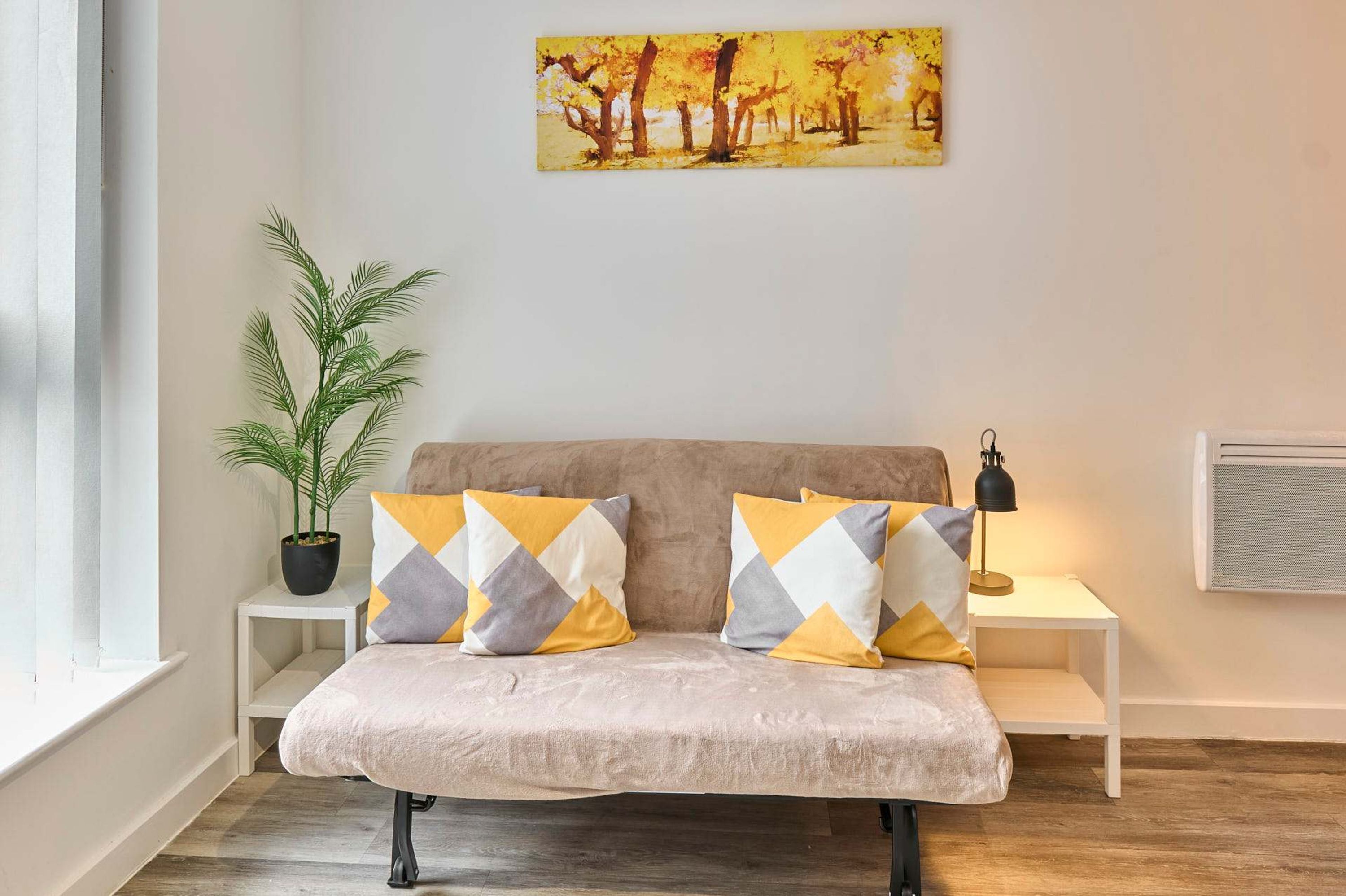 A bed with patterned pillows, a potted plant, a painting, and a lamp on a bedside table.