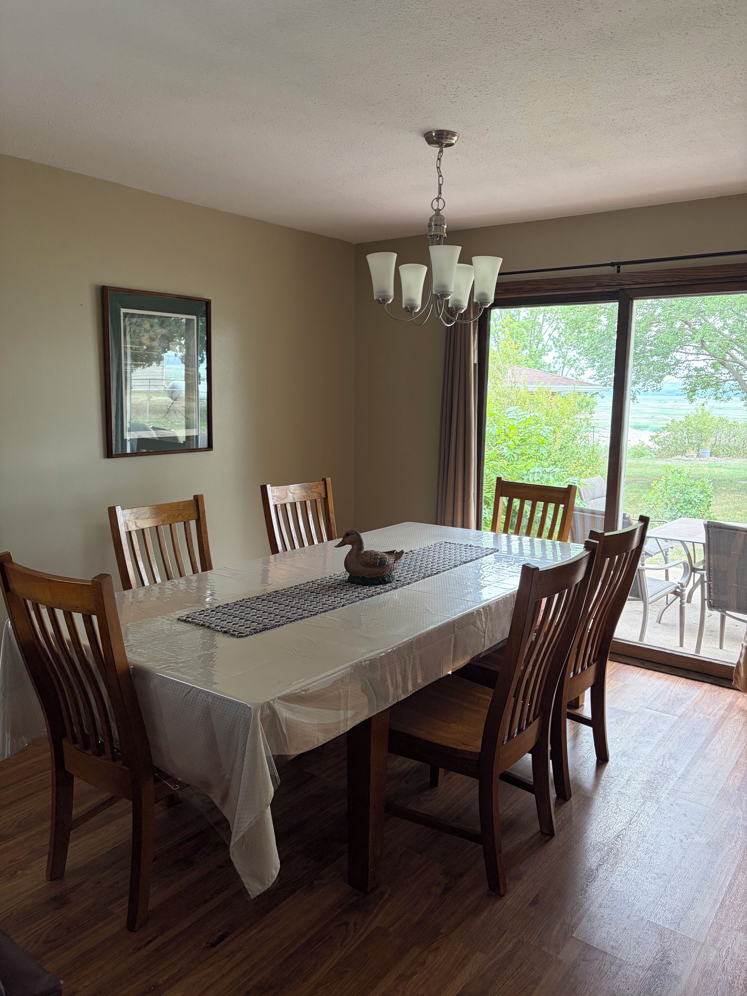 Une salle à manger avec une table dressée pour six personnes, des chaises en bois, un lustre, un tableau encadré au mur et une vue sur l’extérieur par le biais d’une grande fenêtre.