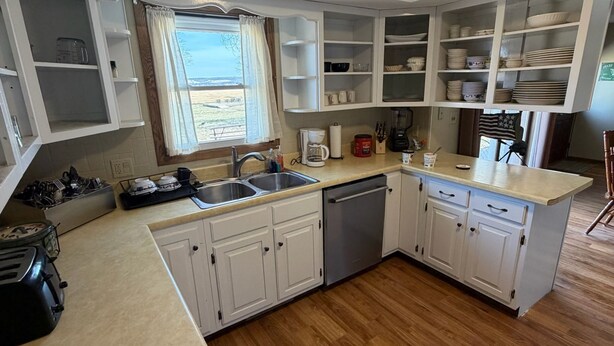 A kitchen with white cabinets, a dishwasher, a sink, and a window with a view of the outdoors. - Bluff’s Bend Retreat (Springfield)