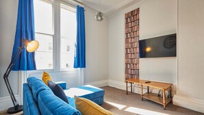 A living room with a blue sofa, a wooden coffee table, a floor lamp, a bookshelf, and a TV mounted on the wall. - Host Stay Flat 2 Club Chambers (Malvern)