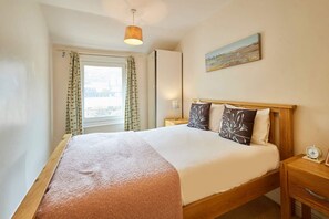 A bedroom with a bed, wooden headboard, bedside tables, a nightstand, a window with curtains, and a painting on the wall. - Host Stay The Beach (Filey)