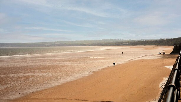 A beach with a wide sandy shore, a calm sea, and a distant hilly landscape.