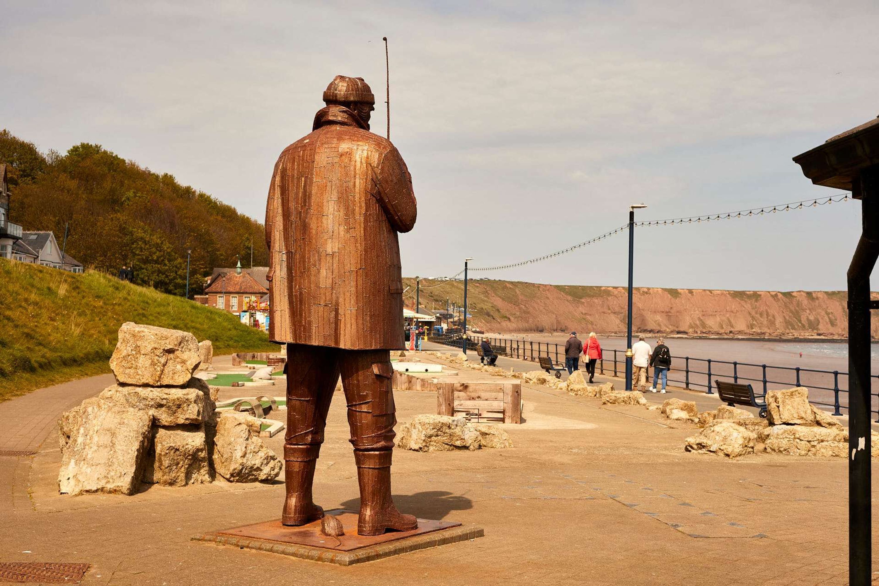 Une promenade en bord de mer agrémentée d’une statue, avec un terrain rocheux et une vue lointaine sur la mer.
