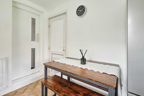 Ein Zimmer mit einer hölzernen Sitzbank, einem weißen Tisch, einer Wanduhr und einer Tür mit Fenster.