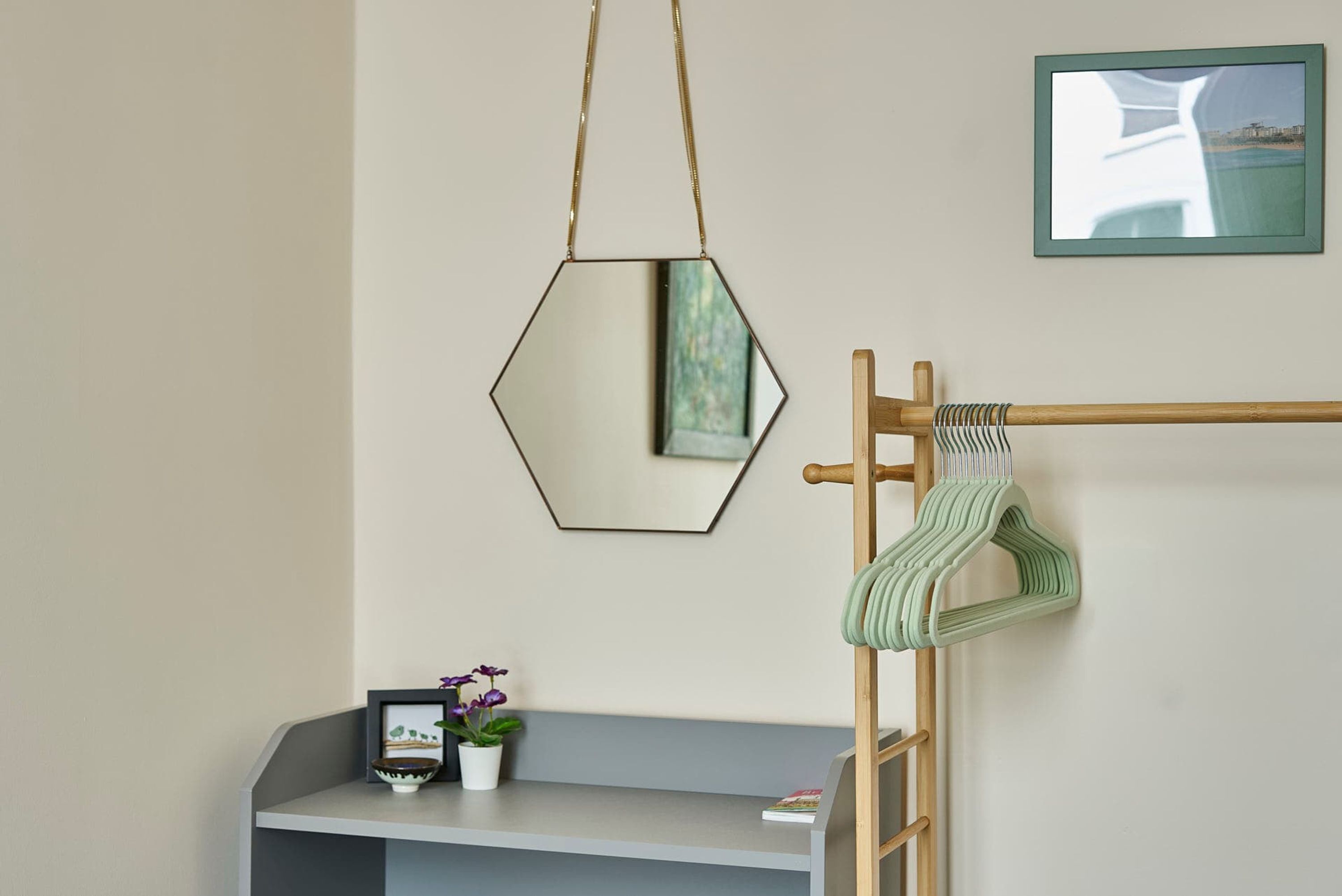 A room with a grey shelf, a hanging mirror, a wooden rack with a green towel, and a framed picture on the wall.