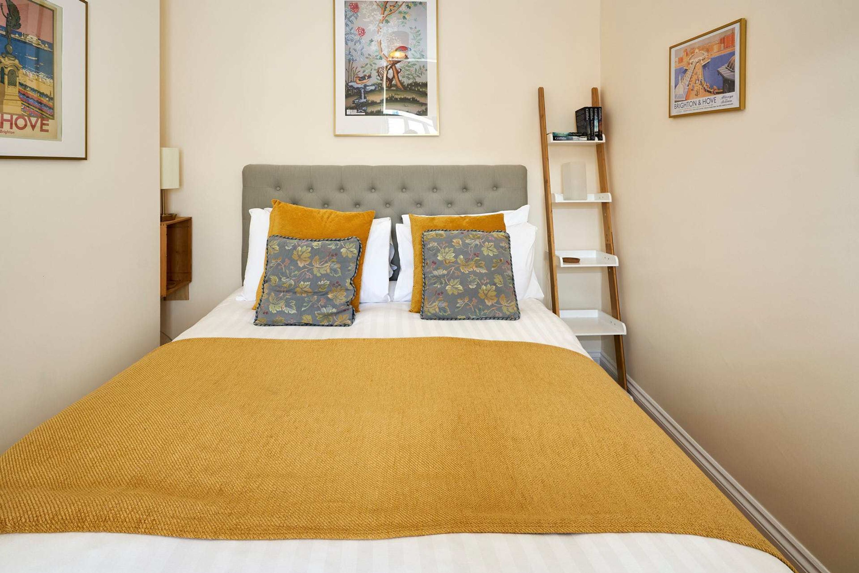 A neatly made bed with a mustard-colored blanket, two floral pillows, and a grey upholstered headboard.