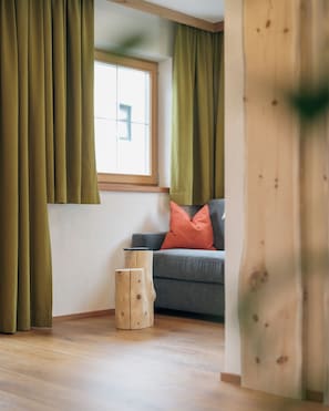 A room with a window, curtains, a sofa, a wooden stool, and a wall with a wooden accent. - Apartment Kreuzspitze (Innervillgraten)