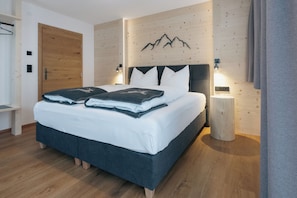 A modern bedroom with a wooden floor, a bed with white bedding, a wooden door, and a wall-mounted light fixture. - Apartment Kreuzspitze (Innervillgraten)