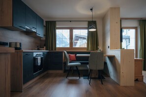A modern kitchen with wooden cabinets, a built-in oven, a dining area with a bench and chairs, and a view of mountains through large windows. - Apartment Kreuzspitze (Innervillgraten)