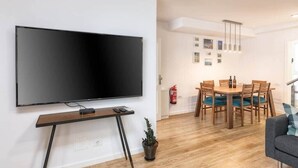 Un soggiorno moderno con una TV a schermo piatto montata a parete, un tavolo da pranzo in legno con sedie, una piccola pianta in vaso e un divano grigio.