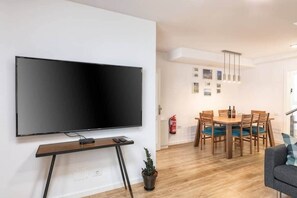A modern living room with a flat-screen TV mounted on the wall, a wooden dining table with chairs, a small potted plant, and a grey sofa. - Tendal Duplex - By Tendal Hosting (Santa Cruz de La Palma)