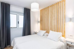 Quarto com cama, cortinas, mesa de cabeceira, luminária e parede de painéis de madeira.