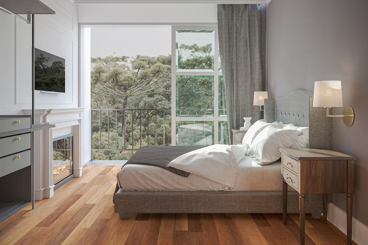 A bedroom with a bed, bedside lamps, a TV, a fireplace, and a balcony with a view of trees.