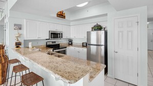 A modern kitchen with white cabinetry, a granite countertop, stainless steel appliances, and a bar area with stools. - Beachfront Bliss, Majestic Sun A1002 | Stunning Gulf Views & Resort Amenities (Miramar Beach)