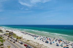 A beach with a road parallel to the shore, lined with beach umbrellas and people. - Beachfront Bliss, Majestic Sun A1002 | Stunning Gulf Views & Resort Amenities (Miramar Beach)