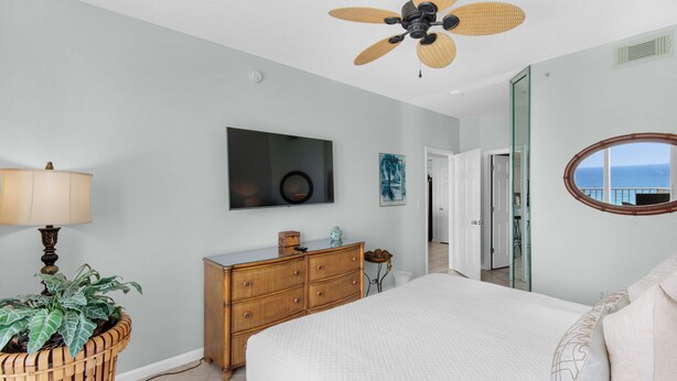 A bedroom with a bed, a dresser, a television, a ceiling fan, and a view of the ocean through a window. - Beachfront Bliss, Majestic Sun A1002 | Stunning Gulf Views & Resort Amenities (Miramar Beach)