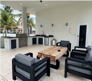 Une cuisine extérieure équipée d’un barbecue, d’un évier et d’un petit réfrigérateur. On y trouve une table en bois avec des chaises et un ventilateur de plafond.