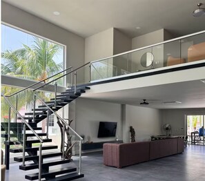A modern interior with a staircase, a flat-screen TV, and a large window with a view of palm trees.