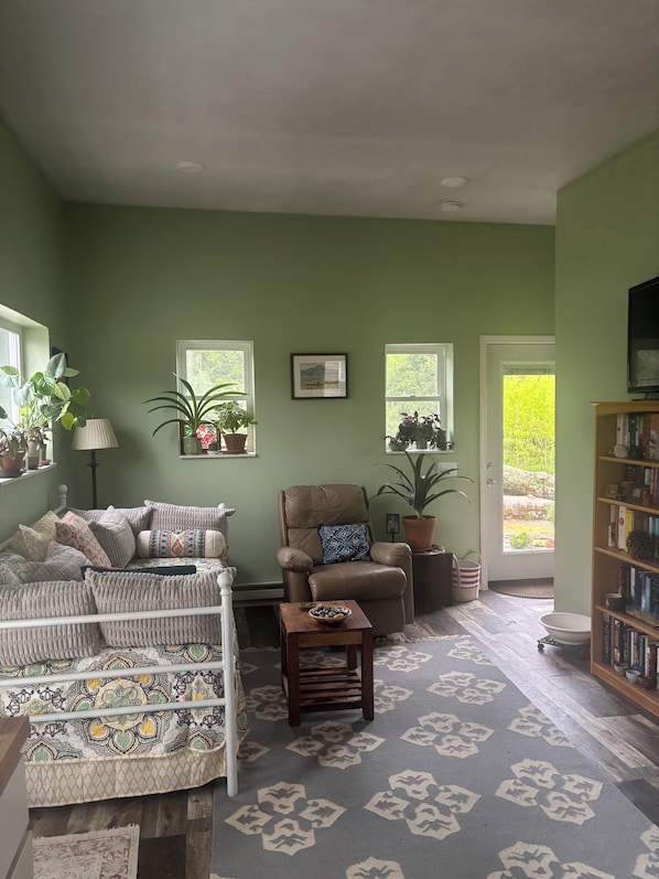 Una sala de estar con sofá, sillón y estantería. Se ven ventanas con plantas y una puerta que da al exterior.